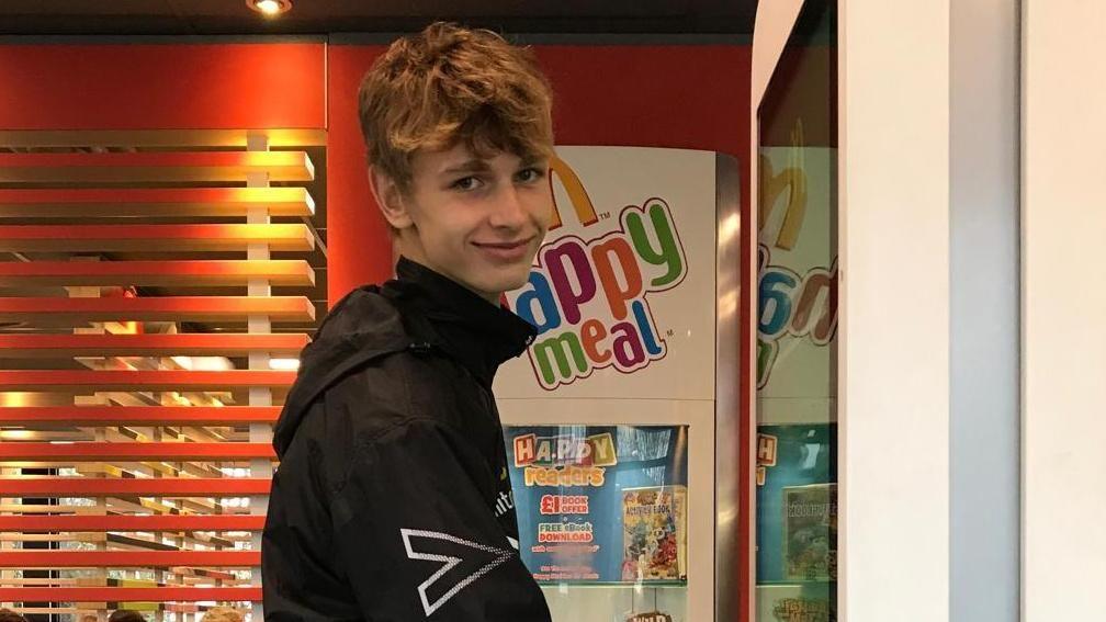 Jacob Crawshaw looking sideways at the camera while standing in front of a fast-food restaurant's self-service ordering board.