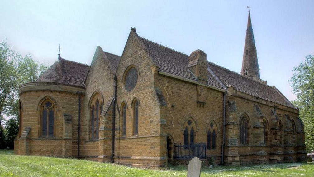 A church building in dark stone in three sections. There are cathedral windows of various sizes and the section nearest the camera also has a round window. There is a spire at the rear of the central section. There is grass around the church and a gravestone is visible.