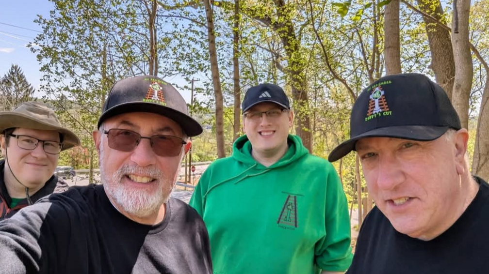 Frank Watson has a grey beard and is wearing a dark long-sleeved top, sunglasses and a Duffy's Cut baseball cap. Bill Watson is clean-shaven and is also wearing a dark top and Duffy's Cut baseball cap. Two other men are also in the picture. Both are wearing glasses, one is wearing a baseball cap and the other a hat.