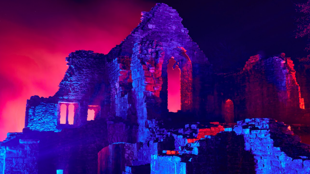 The stone ruins of Finchale Priory, showing remains of stairs, walls and windows, are shining in blue and red as part of the Solitude projection during Durham Lumiere in 2021.