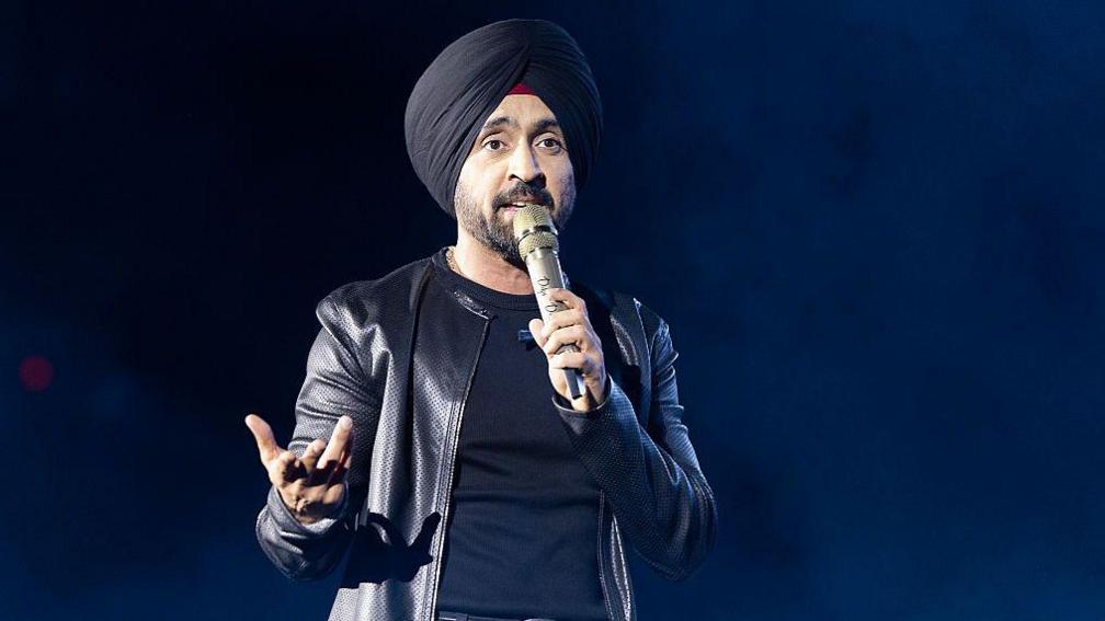Diljit Dosanjh kicks off his Aura World Tour at BC Place on April 23, 2026, in Vancouver, British Columbia, Canada. He is wearing a black jacket over a black t-shirt and sporting a black turban, as he speaks on a microphone from the stage.