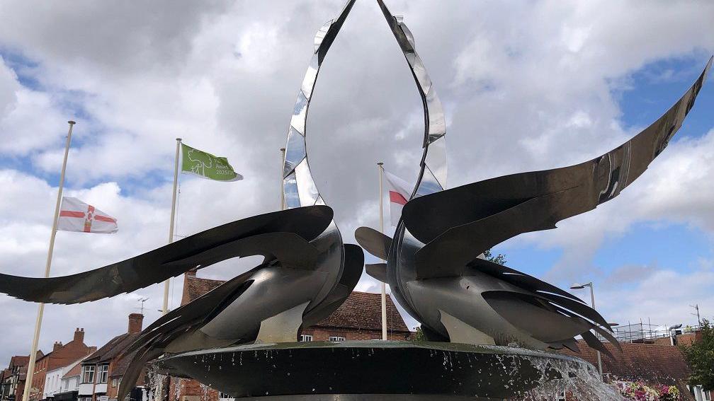 Metal feathers curl out from a central base from which we can see water is falling. Behind them are houses and several flagpoles.