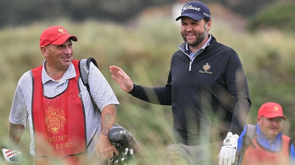 JD Vance, sporting a white golf glove on his left hand, black fleece, blue shirt, grey trousers and blue Titleist cap, is talking to his caddy. The caddy is wearing a red Trump Turnberry cap and tabard, a spotted blue and white shirt, and has heavily tattooed arms. A second caddy, dressed in red hat and tabard and a blue shirt, is in the background. We can make out gorse and heather behind them all.