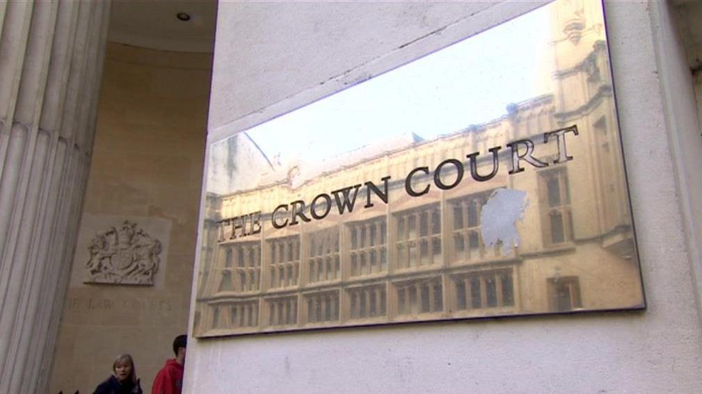 A brass plaque on a sand stone wall reading The Crown Court. A coat of arms is visible on a wall in the background. 