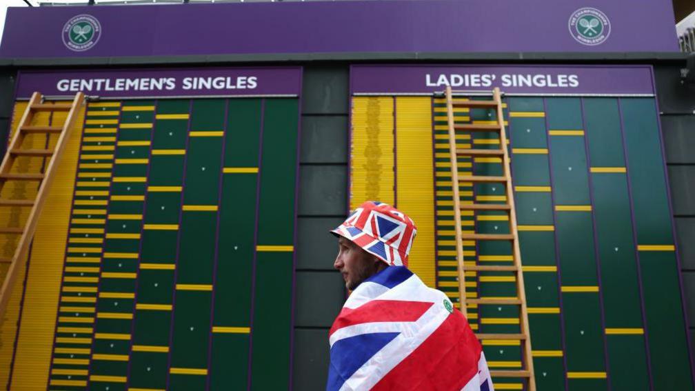 A spectator wearing the Union Flag observes the order of play board during day one of The Championships Wimbledon 2024.