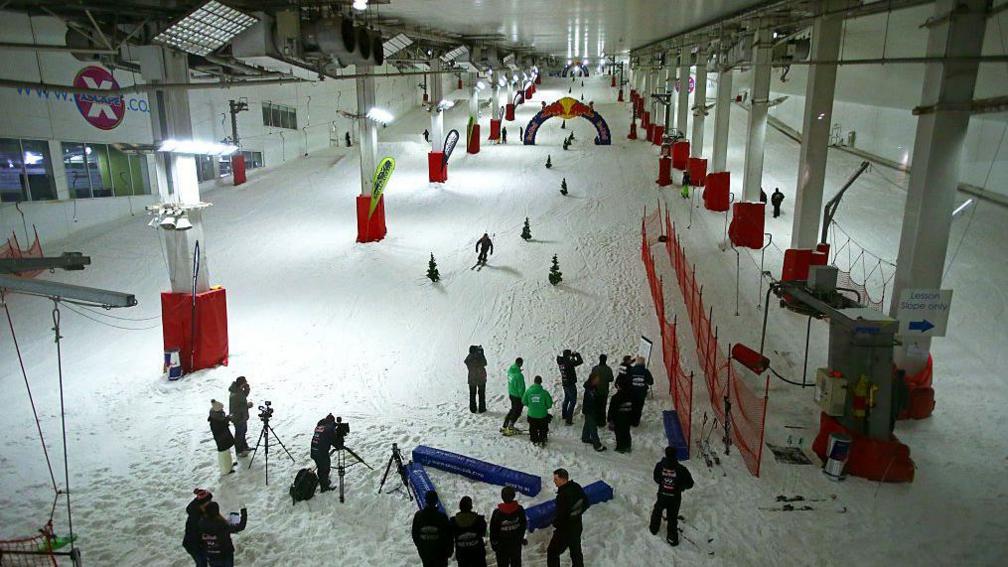 Skiers ski down a long wide ski slops dotted with red crash pads and red fences at the Milton Keynes Snowzone centre.