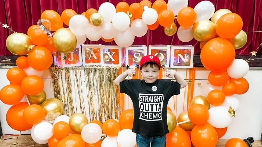 Jaxson standing in a hall in front of a stage with a red velvet curtain, in the middle of a circle of orange, gold and white balloons and some squares spelling 'Jaxson' gold ribbons