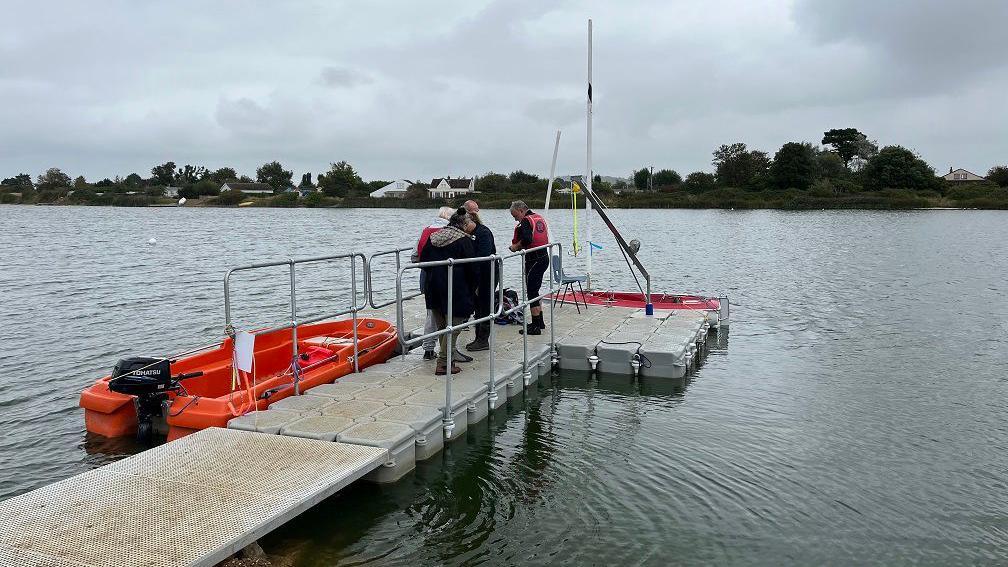 A grey floating pontoon sits on the water. On the pontoon are four people in life jackets getting ready to board a boat at the end of the pontoon. There is a hoist to help people get in. Alongside is a small orange motorboat