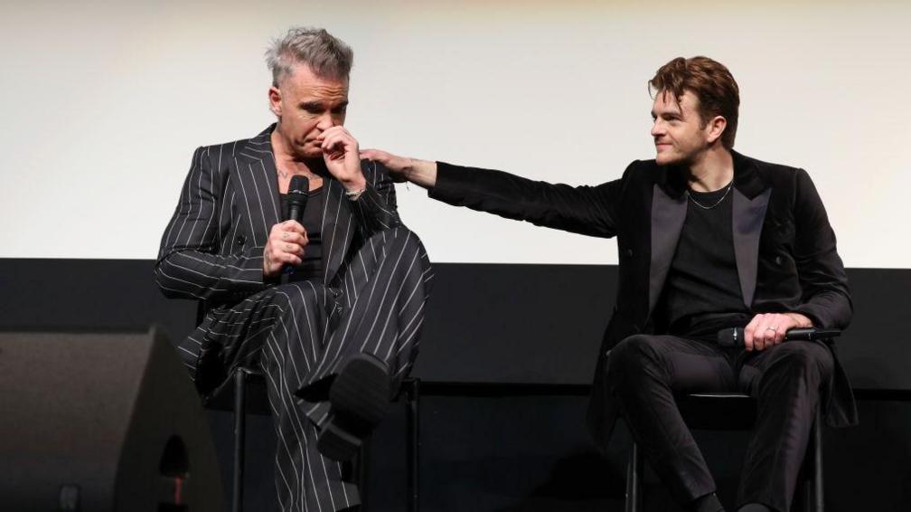 Robbie Williams, wearing a pinstripe suit, is sat on a stage and crying while Jonno puts a reassuring hand on his shoulder.