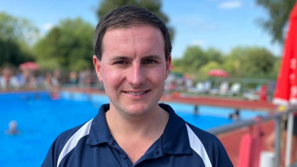 Matt Day is smiling at the camera. He has short, dark hair and is clean-shaven. He is wearing a dark blue polo shirt with white stripes across the shoulders. Behind him is a blurred image of a large swimming pool with sunbeds and parasols