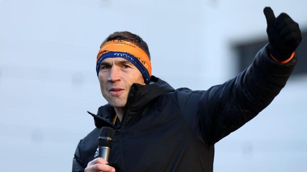 Kevin Sinfield, with short cropped dark hair, holds a microphone and gesticulates, as he wears his fundraising headband and a black coat.