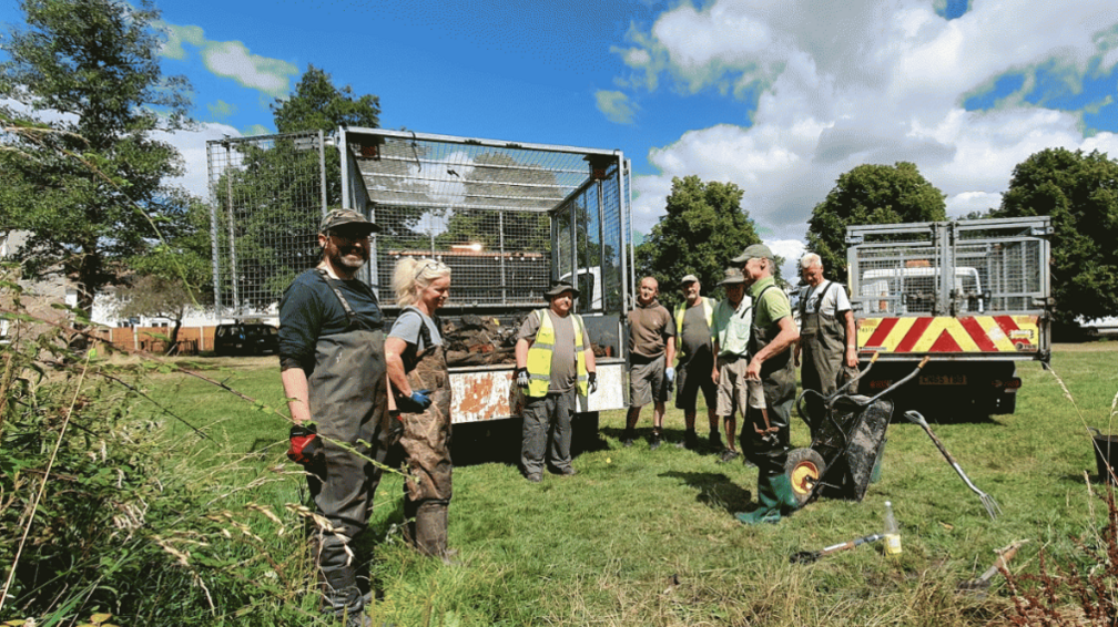 A group of volunteers standing in a field by some council rubbish vans. The are wearing protective dark green clothing and a wheelbarrow in an upright position is near the front of the picture. 