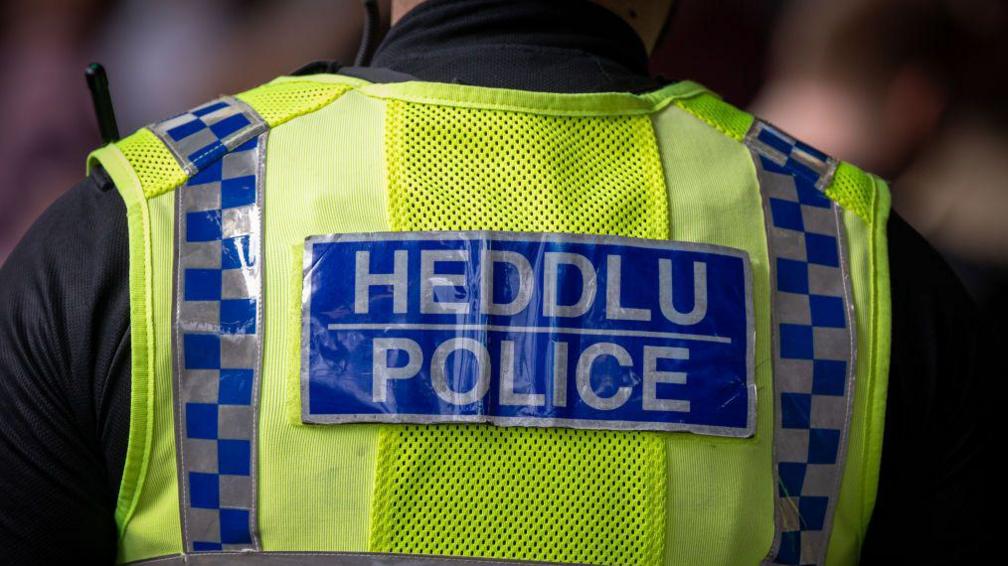 A police officer wearing a fluorescent yellow vest with "Heddlu" and "Police" written on the back. 