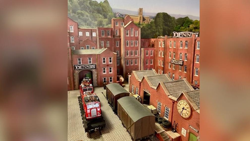 A close up of a model which shows the red brick buildings of the Dennis factory along with signage and a model fire engine. In the background Guildford Cathedral is seen on a hill along with trees.