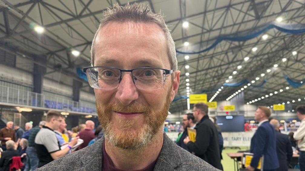Philip McGuigan has short brown hair which is spikey at the front and a short ginger beard. He is wearing glasses and a tweed grey jacket and is standing in an election count centre.
