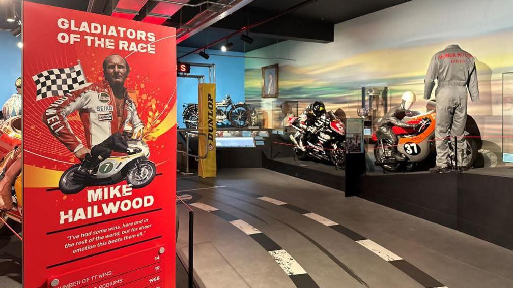 The Isle of Man TT gallery featuring classic motorbikes from the races and a set of overalls. A red information board with the title Gladiators of the Races and featuring a picture of Mike Hailwood is in the foreground.