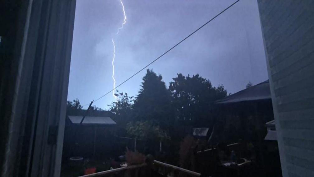 An image of lightning taken through a window illuminating a garden with sheds and trees.