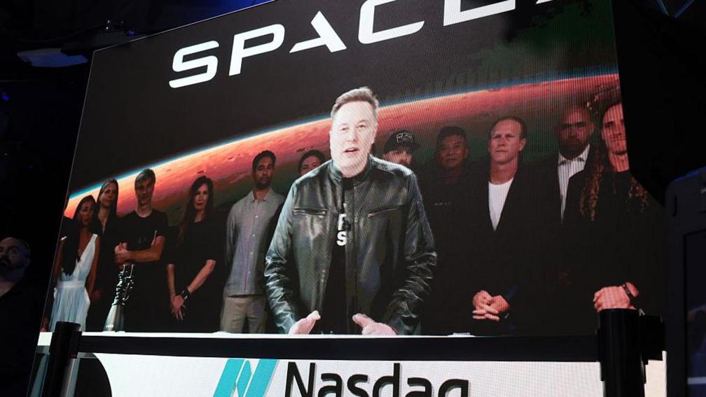 Elon Musk stands at a podium with the Nasdaq logo on it wearing a black t shirt and black leather jacket. 