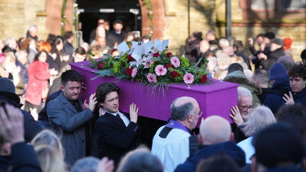 The pink glittery coffin of Linda Nolan is carried out of St Paul’s Church. It has pink and red flowers on top. Hundreds of people are watching.