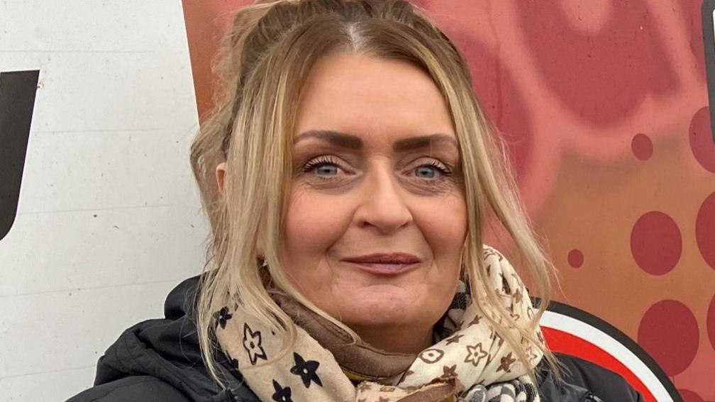 A woman with shoulder length blonde hair is looking at the camera. Only her head and shoulders are visible. She appears to be outside, standing in front of an image but the picture is too close-up to make out what it is. The image is white and red. She is wearing a scarf and a black coat. 