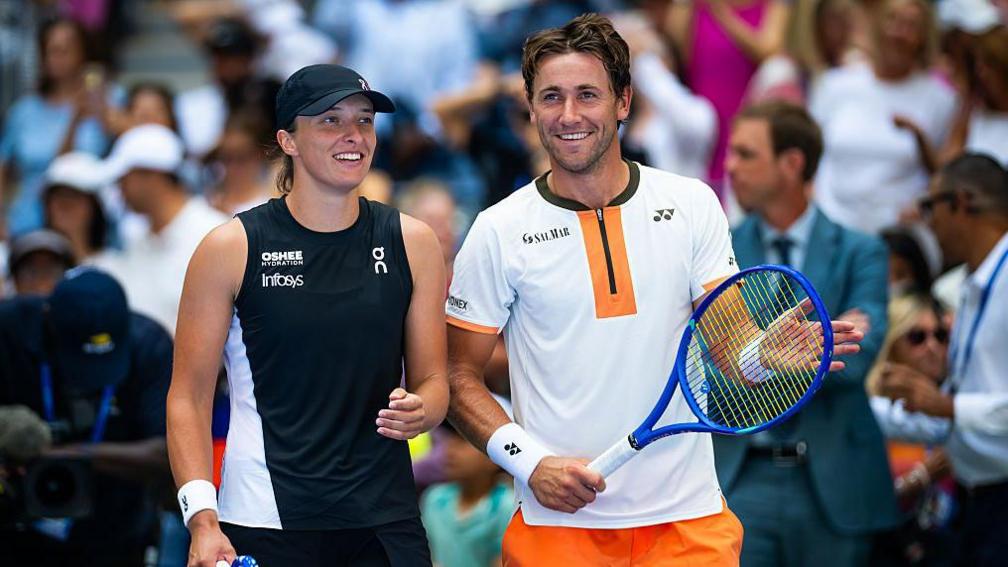 Iga Swiatek and Casper Ruud smile while in doubles action together at the US Open, with Swiatek in a mainly black sleeveless tennis top and dark cap, while Ruud wears a largely white top with black and orange trim and holds a blue racquet, with spectators in the background.
