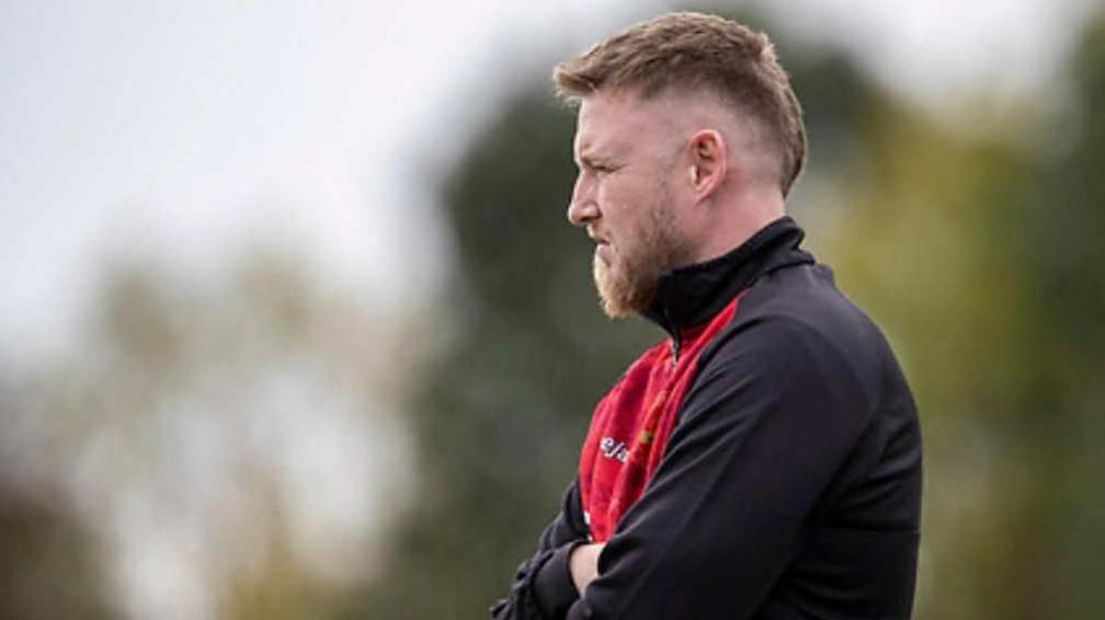 A blond-haired man pictured in profile, with his arms folded wearing his football coach jacket.