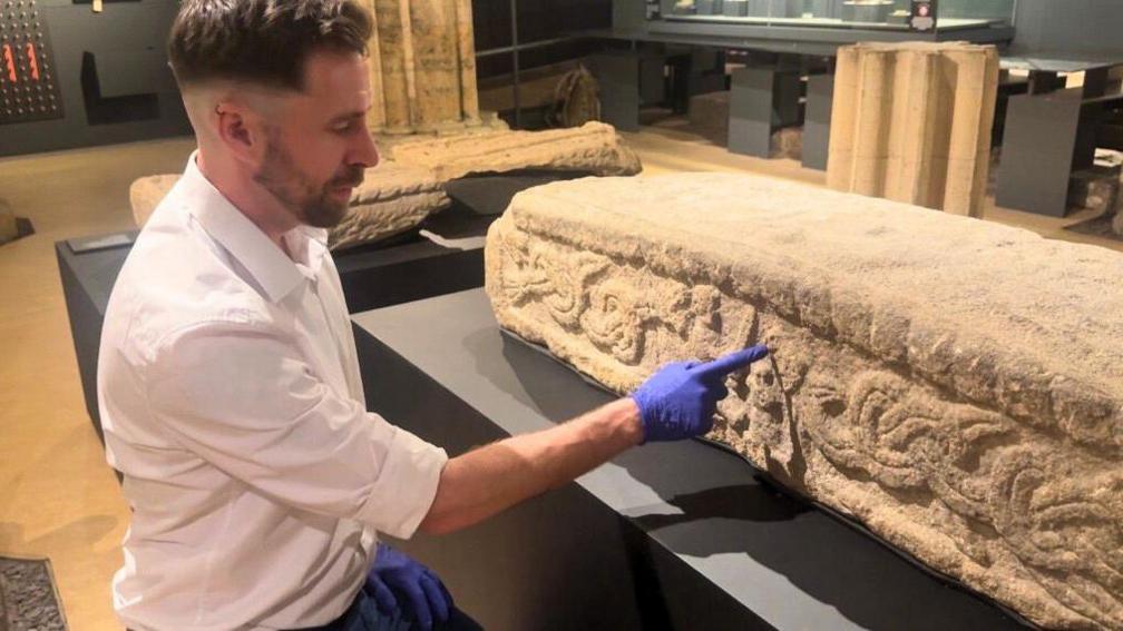 A man in a white shirt and blue gloves points to carvings in stonework.