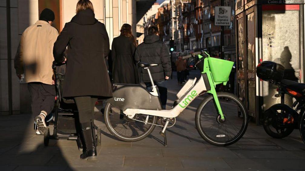 A Lime rental e-bike is left on a busy London pavement as people walk past.