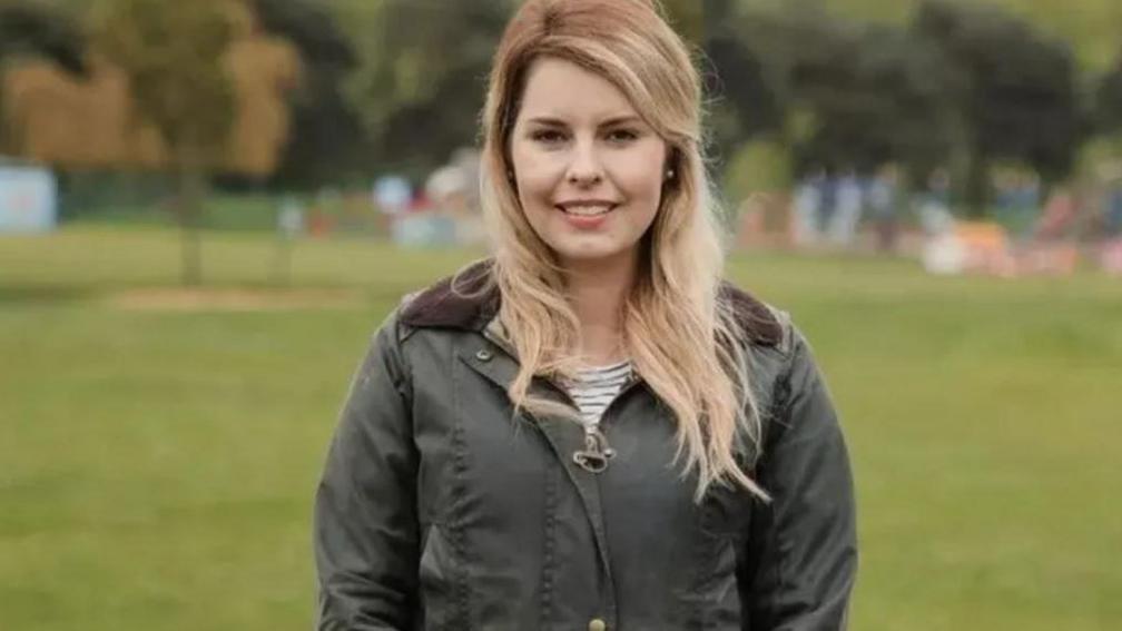 Kim McGuiness smiling directly at the camera. She is standing in a field and wearing a green waterproof jacket.