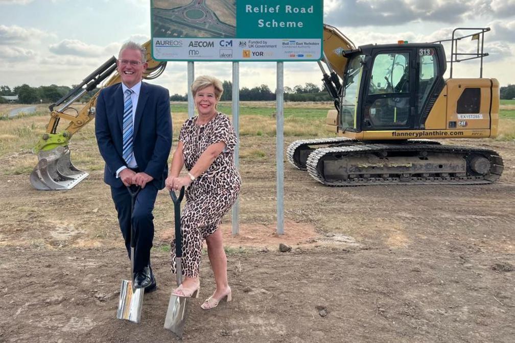 A man in a black suit is smiling and holding a spade with one foot rested on it. A woman next to him with short blonde hair and a leopard print dress is also smiling and has one foot on a spade, she is wearing beige shoes with small heels. A sign can be seen stating 'Howden Relief Road Scheme' in the background as well as a digger.