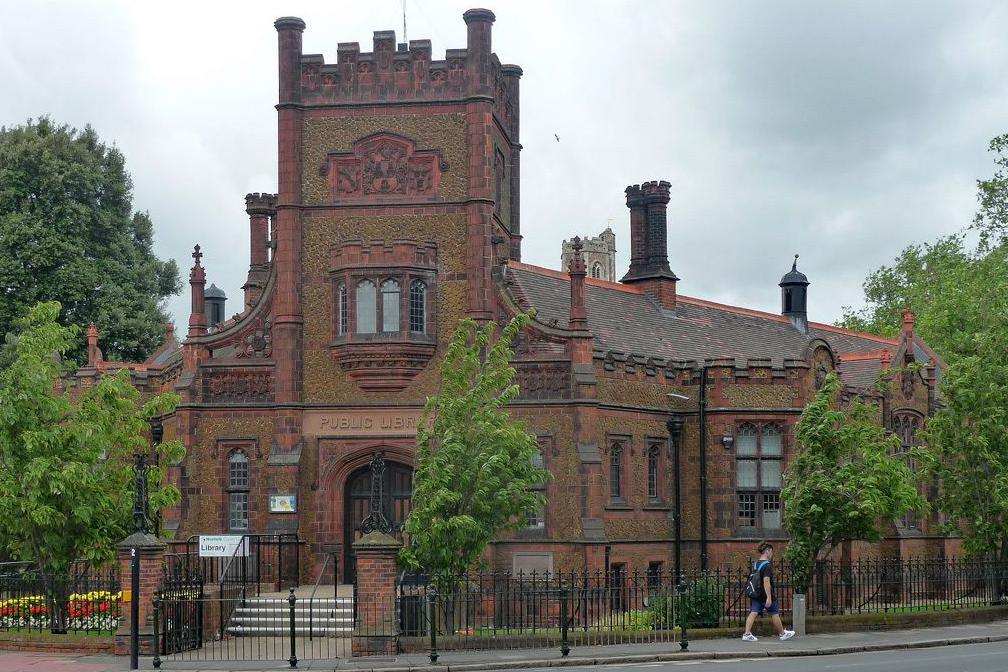 The ornate brick, turretted Carnegie Library on London Road in King's Lynn. The entrance has a tower on top of it, with the building lined with small trees, flowers and iron railings