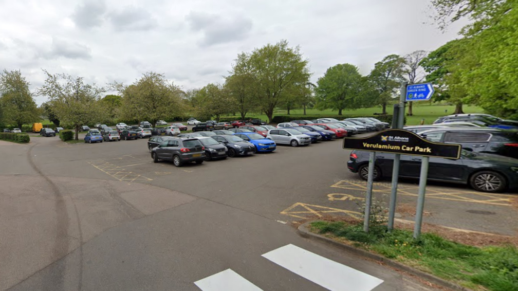 Rows of cars parked in a surface car park with trees and an open green space surrounding it.