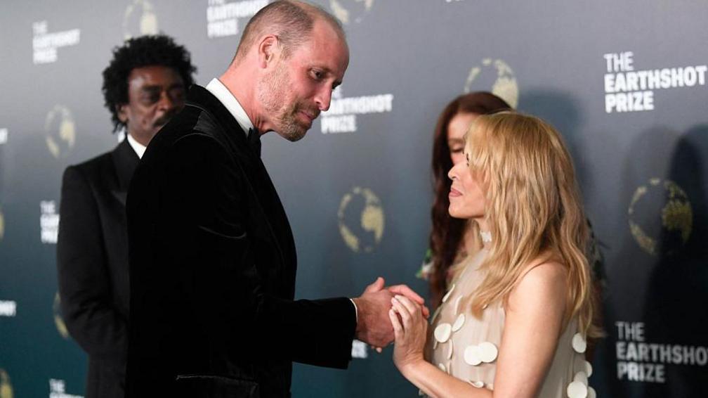 Prince William smiles and wears a black velvet jacket as he touches hands with a Kylie Minogue, who is wearing a beige dress with white disc adornments