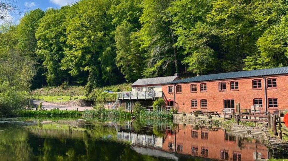 An old industrial mill-like building, surrounded by trees, overlooks a pond. A road can be seen leading up to the building, which has outdoor seating. It is a sunny day.