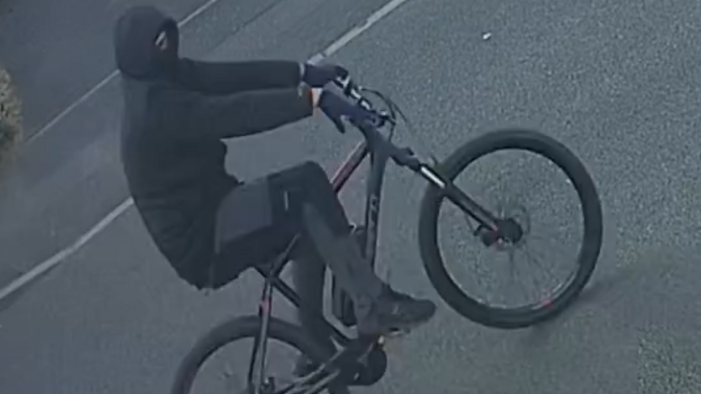 A man on a black bicycle who is dressed completely in black, including gloves and a balaclava