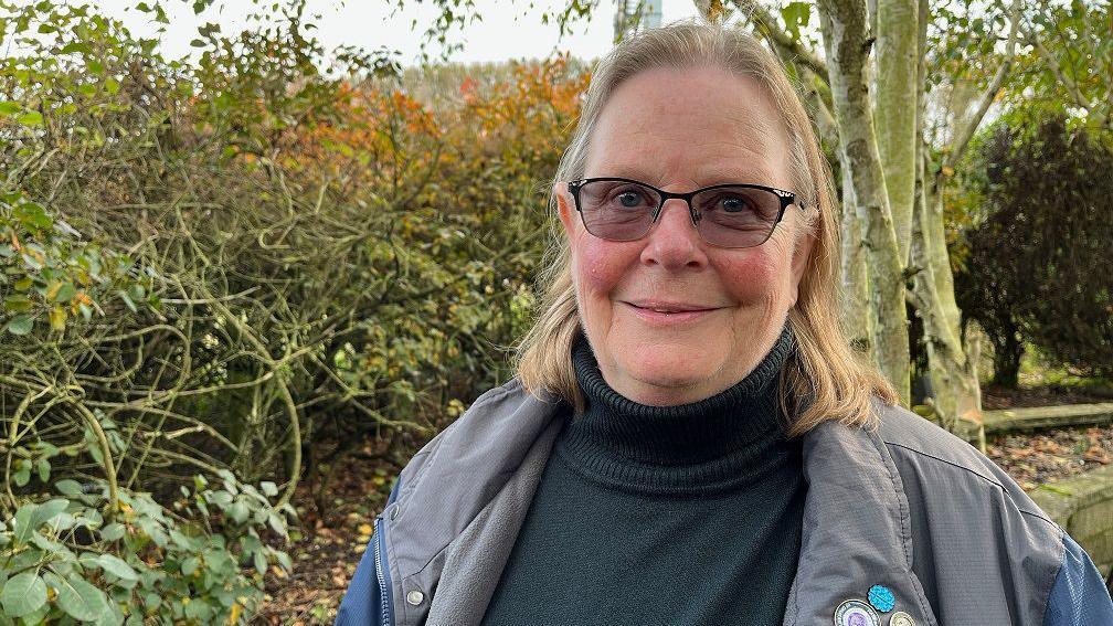 A woman with short light blonde hair wearing glasses, a black polo neck and a light grey waterproof jacket, smiling at the camera with trees and shrubbery behind her.