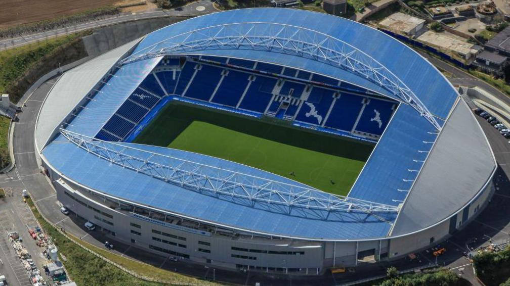 An aerial shot of the Amex Brighton and Hove Albion football ground at Falmer, East Sussex. The blue curved roof encloses the green pitch.