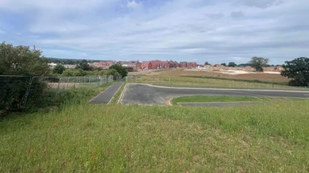 A field with an unfinished road running through and across it. In the distance is a housing estate.
