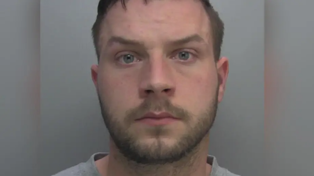 A police custody photo of Shaun Dyson, who stares at the camera. He has short brown hair and a beard and wears a grey collarless top.