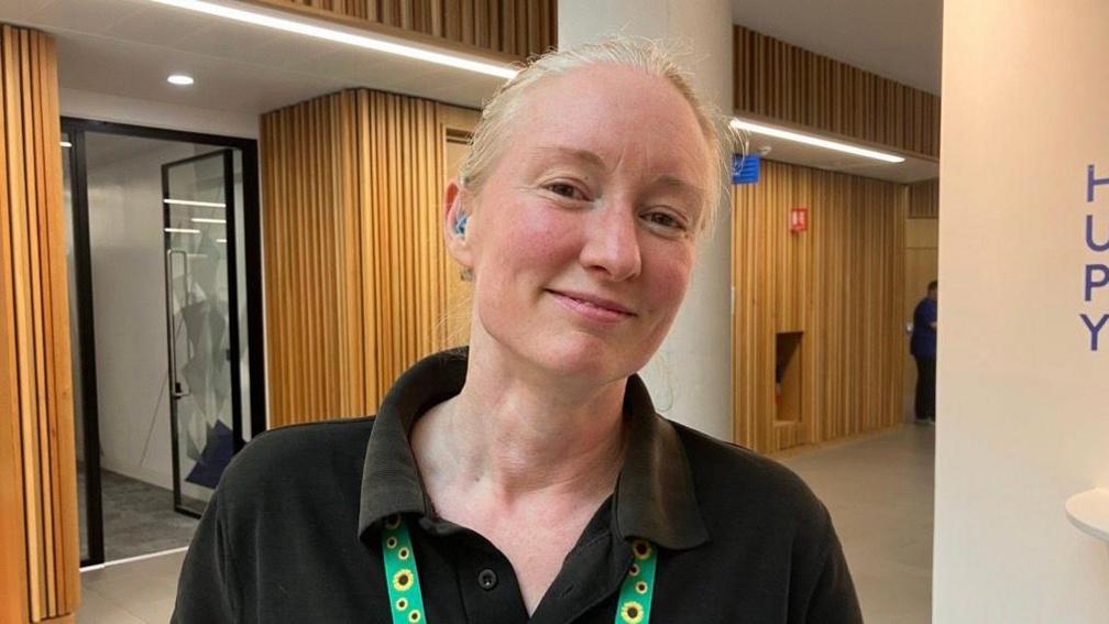 A woman with white blonde hair smiling at the camera. She is wearing a black polo shirt, a sunflower lanyard and has a blue hearing aid.