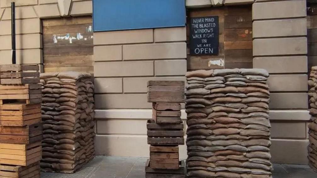 Sandbags are stacked next to a building in Hull's Old Town. On it is a sign stating: "Never mind the blasted windows. Walk right in. We are open."