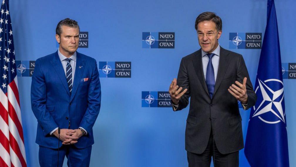 Pete Hegseth stands still in a blue suit in front of a blue Nato backdrop while Mark Rutte talks