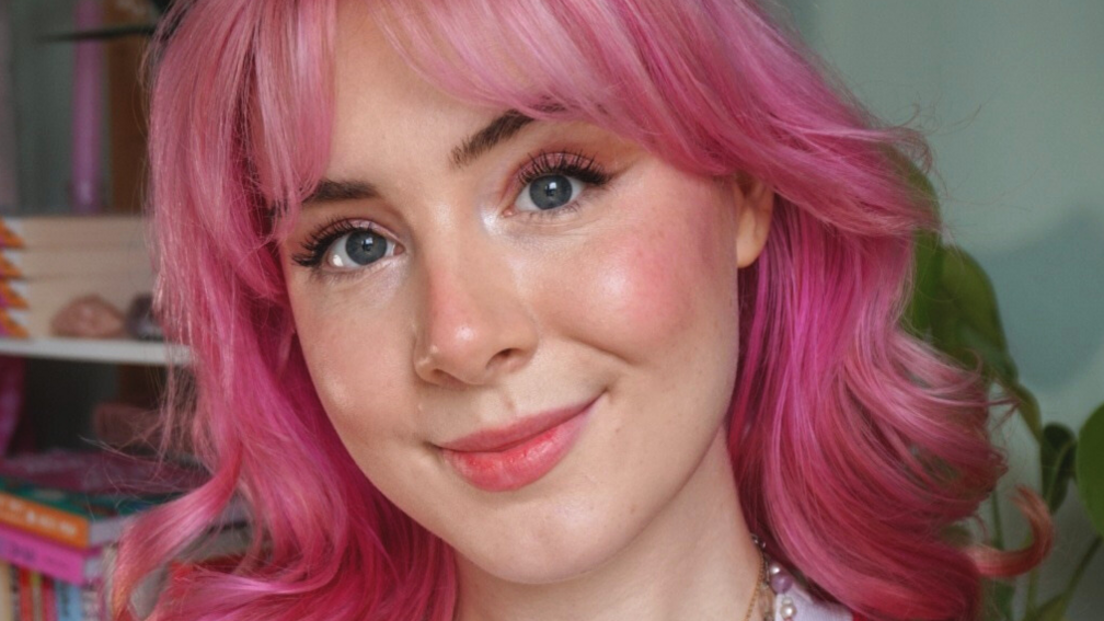 Milly Evans who has pink wavy shoulder-length hair and is standing in front of a bookcase. 