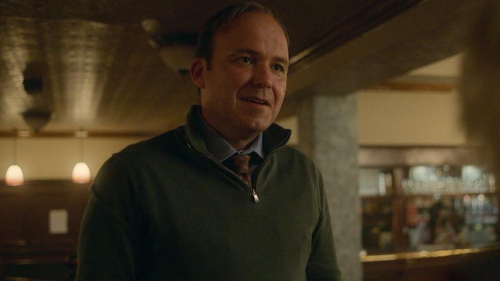 Rory Kinnear in a scene in Toxic Town. He is wearing a zipped jumper and a shirt with a tie. He is standing inside a pub and smiling.