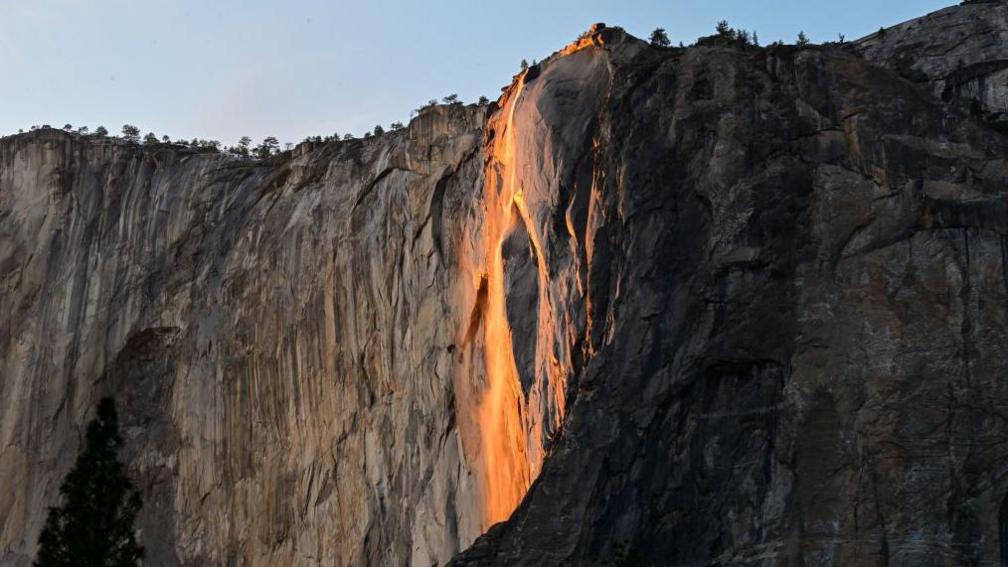 waterfall down the side of a mountain has an orange glow due to the sunset.