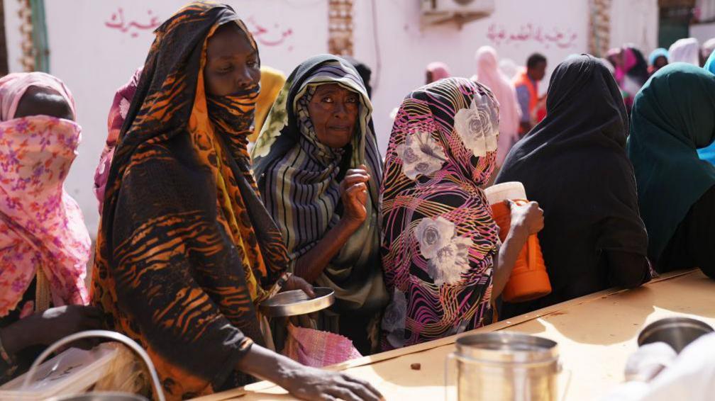 Women in colourful headscarves wait in a queue for food to be distributed.