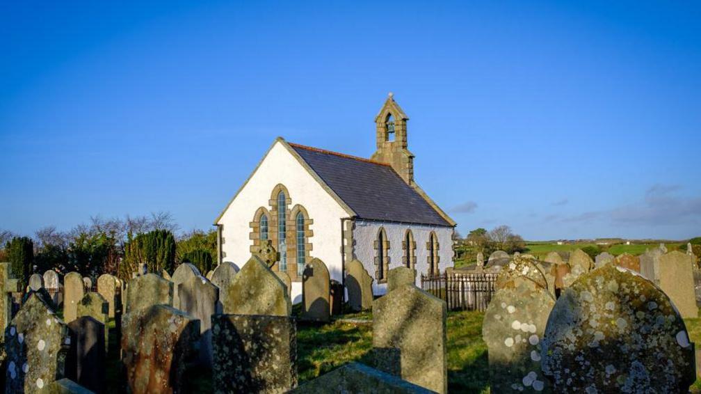 A large number of grave stones around an old white Church, there are fields in the background.