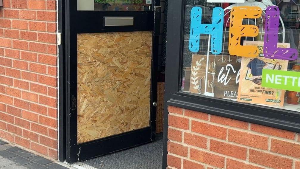 A closer view of the door of the charity shop. A wooden panel has replaced the glass after it was smashed. The panel is situated in the lower part of the door.