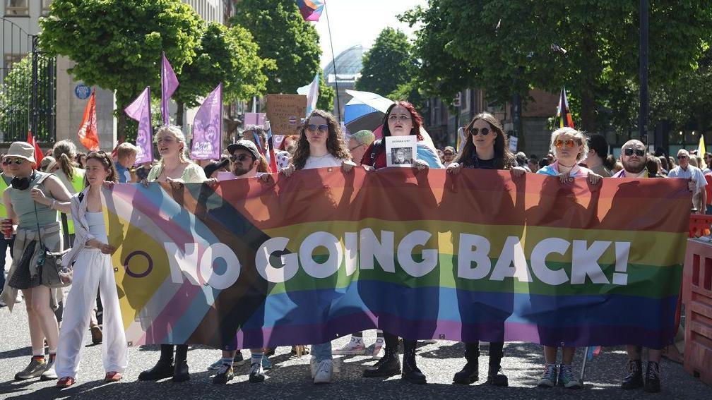A line of eight people at the front of a protest march are holding a banner with the words 'No going back!' The banner features the colours of the transgender flag and pride flag