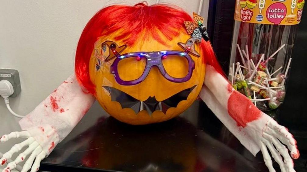 A pumpkin wearing a red wig, purple glasses and a black bat for a mouth. It has two skeleton arms sticking out either side of it. There is a plastic tube of lollipops in the background.